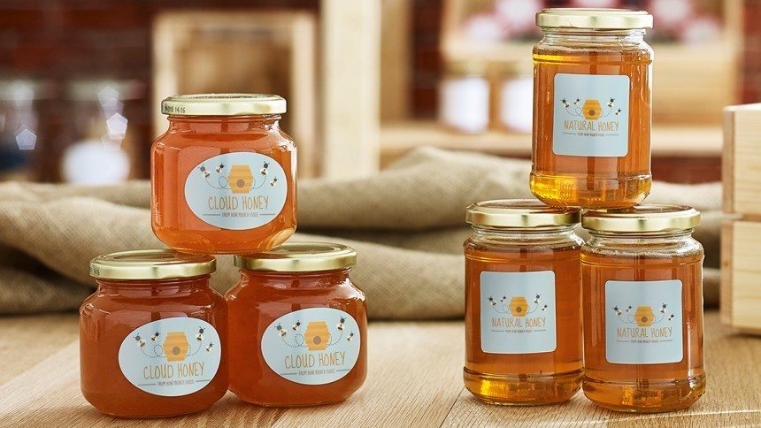 Mai multe borcane cu miere și capace aurii, aranjate pe o masă de lemn. Unele etichete poartă inscripția "Cloud Honey", iar altele "Natural Honey".