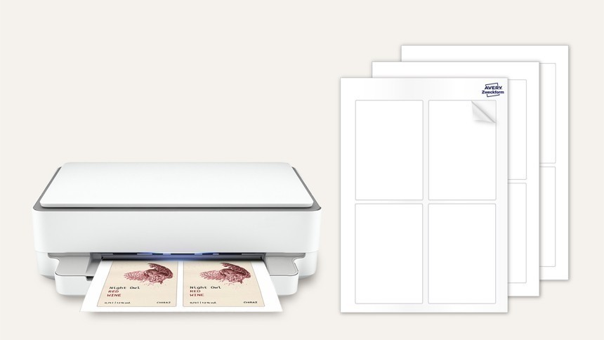 O imprimantă albă imprimă o coală cu două etichete pentru vin cu un design de bufniță. Alături se află trei coli goale de etichete autoadezive Avery.