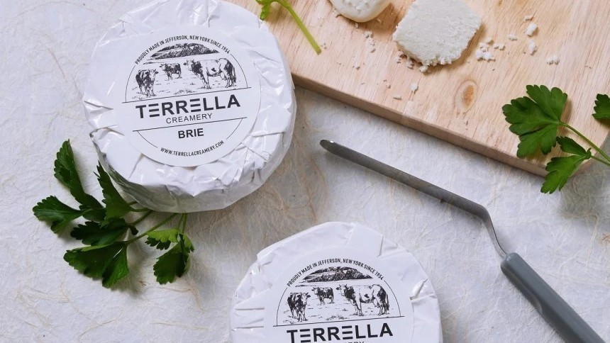 Două brânzeturi rotunde de brie în ambalaj alb cu eticheta "Terrella Creamery", așezate pe o masă lângă un tocător și un cuțit de brânză.