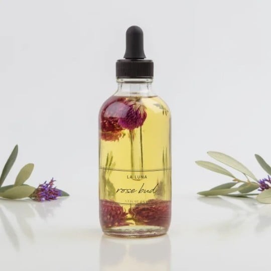 O sticlă transparentă cu un picurător negru, umplută cu ulei în care sunt scufundați boboci de trandafir uscați. Eticheta poartă numele "La Luna rose bud". În fundal se văd crenguțe de măslin.