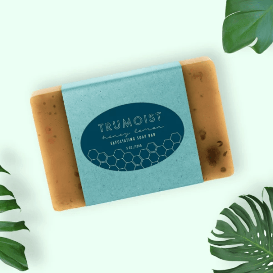 Un săpun solid exfoliant dreptunghiular, cu miere și lămâie, "Trumoist". Săpunul este învelit într-o etichetă de hârtie albastră cu un motiv de fagure de miere. Acesta este așezat pe un fundal verde deschis, cu frunze de monstera în colțurile imaginii.