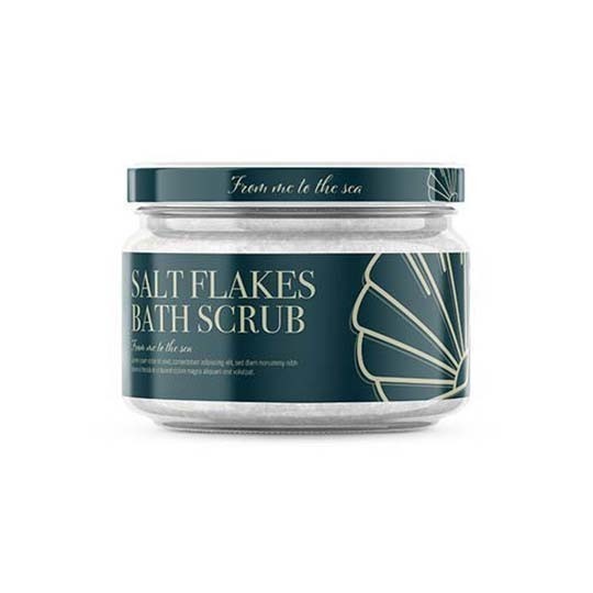 Un borcan de sticlă cu scrub de baie "Salt Flakes Bath Scrub". Eticheta de culoare verde închis este decorată cu o grafică stilizată a unei scoici, iar capacul are aceeași culoare. Fundalul este alb.