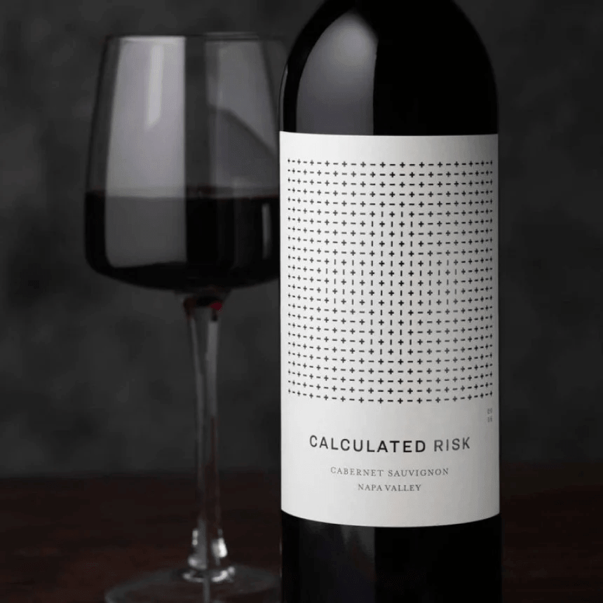 O sticlă de vin roșu "Calculated Risk" cu o etichetă elegantă, minimalistă, având un model format din semne plus și minus, așezată lângă un pahar de vin.