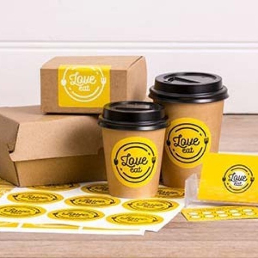 Pahare de cafea din hârtie și o cutie de carton pentru take-away, cu etichete rotunde, galbene, cu logo-ul "Love Eat". Pe masă se află o coală cu aceleași etichete.