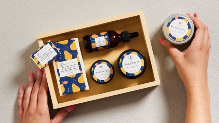 O tavă din lemn cu un set de produse cosmetice marca "Pearberry". Ambalajele au un model bleumarin cu ilustrații de pere galbene. Două mâini țin o cutie metalică rotundă din aceeași serie.