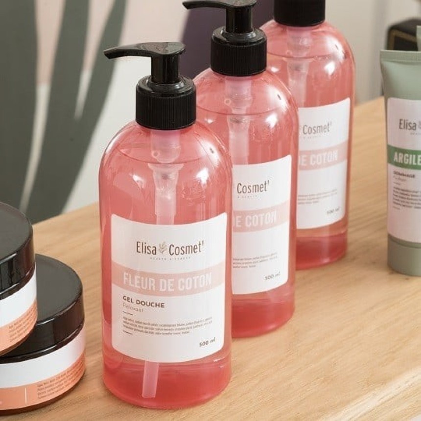 Trei sticle transparente cu pompiță neagră, umplute cu gel de duș roz "Fleur de Coton" de la Elisa Cosmet'. Acestea sunt așezate pe o poliță de lemn, înconjurate de alte produse cosmetice.