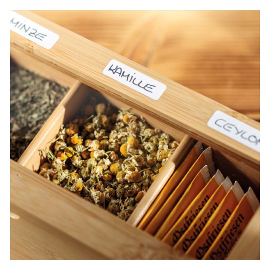 Imaginea descrie o cutie de lemn care conține diverse ceaiuri din plante. Aceasta arată compartimente etichetate cu diferite tipuri de ceaiuri, inclusiv mușețel (kamille) și Ceylon, cu flori uscate de mușețel vizibile într-unul dintre locuri.