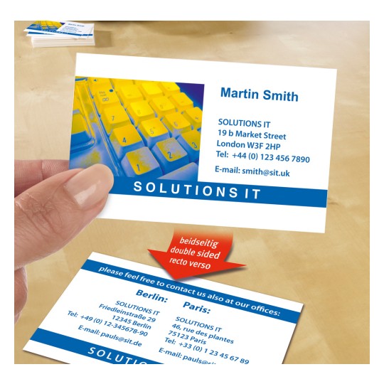 Imaginea arată o carte de vizită ținută între degete, pe care sunt afișate detalii de contact și informații pe două fețe pentru o companie numită 'SOLUTIONS IT'. În fundal este parțial vizibilă o tastatură colorată.