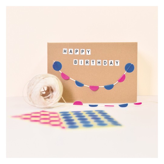 Imaginea prezintă un card de ziua de naștere cu cuvintele "LA MULȚI ANI" afișate pe el. Cardul este decorat cu un șir de cercuri albastre și roz. În apropiere, se află un mosor de sfoară și o hârtie imprimată cu puncte colorate.