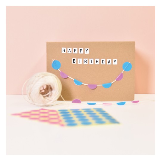 Imaginea prezintă o felicitare maro cu inscripția 'LA MULȚI ANI' scrisă în litere. Există o ghirlandă decorativă din cercuri albastre și mov agățată de-a lungul felicitării. În prim-plan se află un ghem de sfoară, împreună cu hârtii cu modele în roz, galben și albastru.