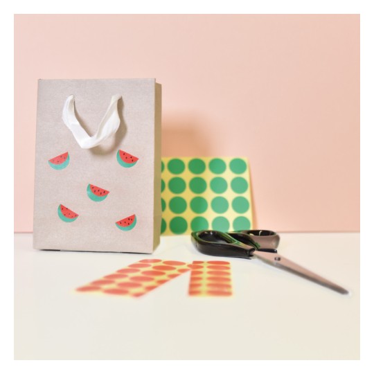 Imaginea prezintă o plasă de cadouri decorativă din hârtie, împodobită cu autocolante cu pepene verde. Aceasta este însoțită de hârtie cu modele, o pereche de foarfece și autocolante rotunde și colorate, așezate pe o suprafață plată cu un fundal roz pal.