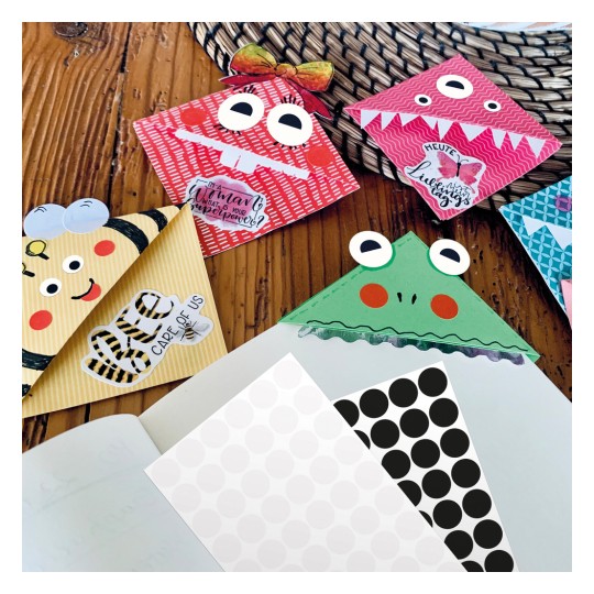 Imaginea prezintă o colecție de modele de plicuri colorate și fanteziste care seamănă cu diverse personaje, inclusiv un monstru, o albină și o broască. Acestea sunt așezate pe o suprafață din lemn alături de hârtie cu modele și un caiet gol.