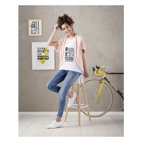 Imaginea prezintă o tânără cu păr creț așezată pe un taburet, purtând un tricou alb cu un mesaj, un cardigan roz deschis și blugi albaștri. În fundal, sunt două imprimeuri înrămate și o bicicletă.