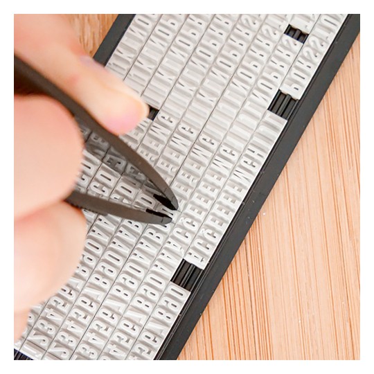 Imaginea arată o mână folosind o pensetă pentru a selecta litere dintr-un set de plăci mici de plastic alb aranjate în rânduri, cu o suprafață de lemn dedesubt.