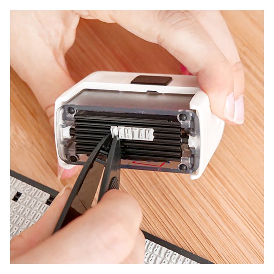 Imaginea arată o persoană folosind o pensetă pentru a manipula un obiect mic în interiorul unui dispozitiv de mână sau ștampilă. Fundalul prezintă o suprafață de lemn cu ștampile sau litere suplimentare vizibile.