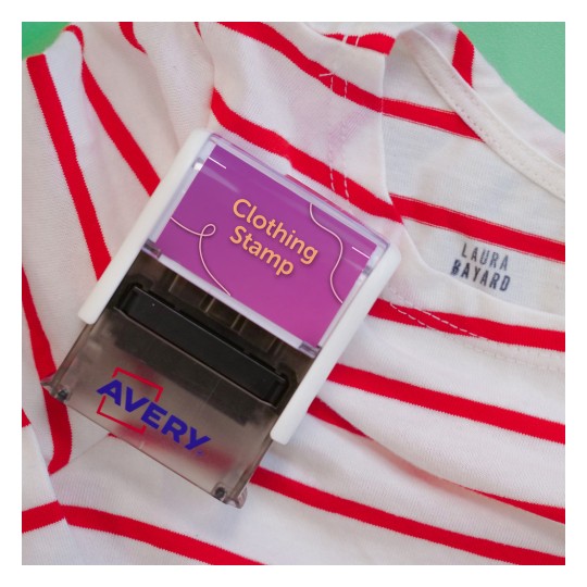 Imaginea arată o ștampilă pentru haine, marcată 'Clothing Stamp', plasată pe o cămașă cu dungi roșii. Cămașa are o etichetă pe care scrie 'LAURA BAYARD'. Ștampila este de la brandul 'Avery'.