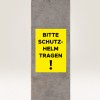 Imaginea prezintă un semn galben pe un stâlp de beton, pe care scrie 'BITTE SCHUTZ-HELM TRAGEN!', ceea ce se traduce prin 'Vă rugăm să purtați o cască!', indicând o cerință de siguranță.