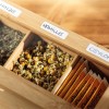 Imaginea descrie o cutie de lemn care conține diverse ceaiuri din plante. Aceasta arată compartimente etichetate cu diferite tipuri de ceaiuri, inclusiv mușețel (kamille) și Ceylon, cu flori uscate de mușețel vizibile într-unul dintre locuri.